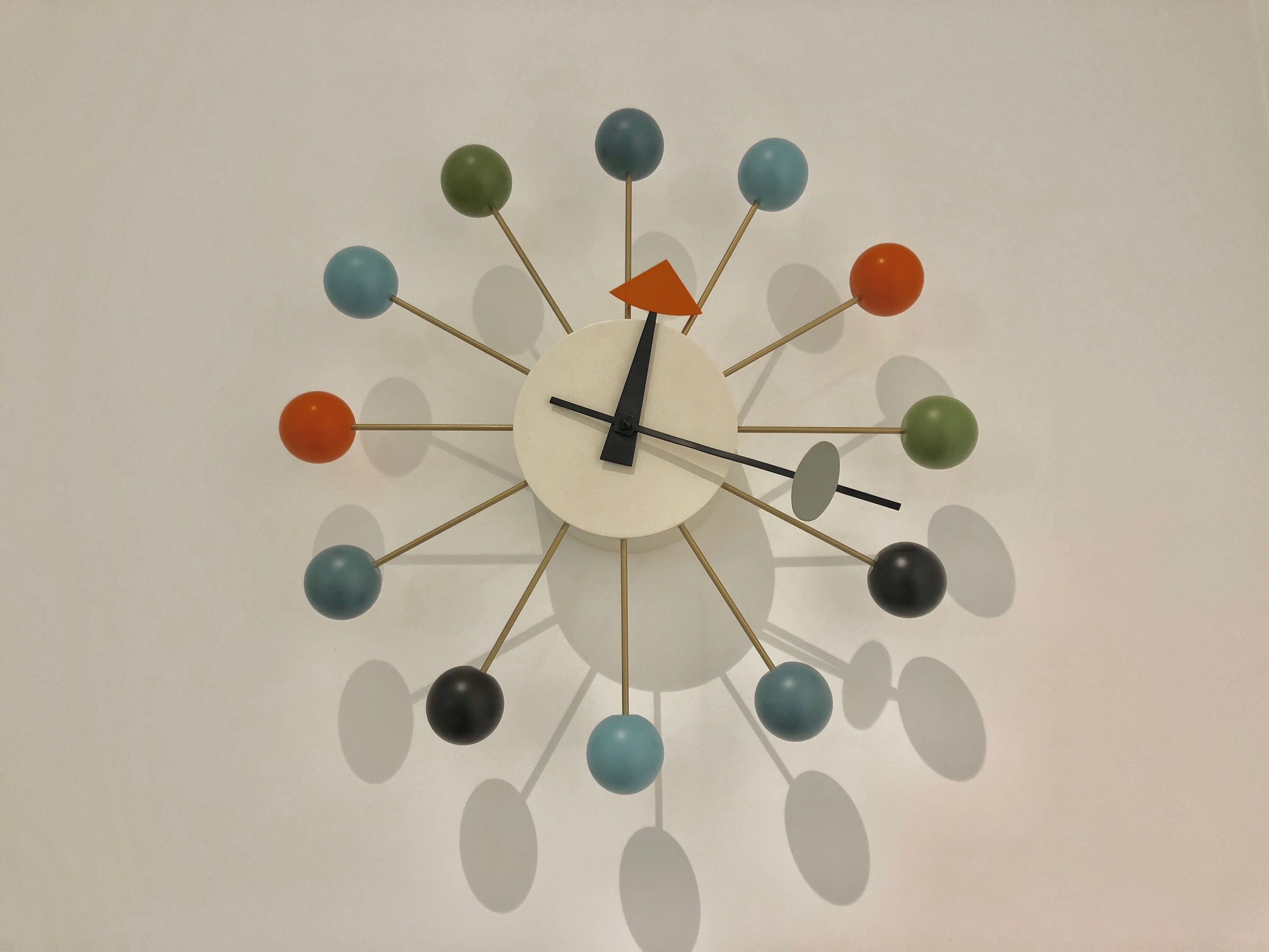 Un reloj de pared con diseño minimalista, que presenta una esfera blanca central y múltiples indicadores de hora de colores.