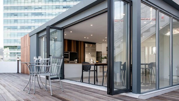Un bâtiment moderne à la façade vitrée, doté d’une terrasse en bois et d’un espace repas avec des chaises et une table.