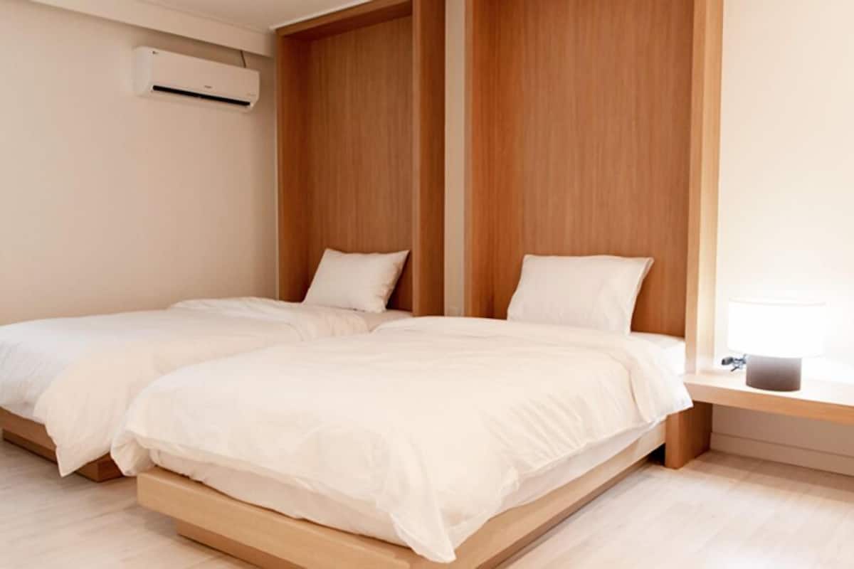 A bedroom with two single beds, a wooden headboard, a bedside table with a lamp, and an air conditioning unit on the wall.