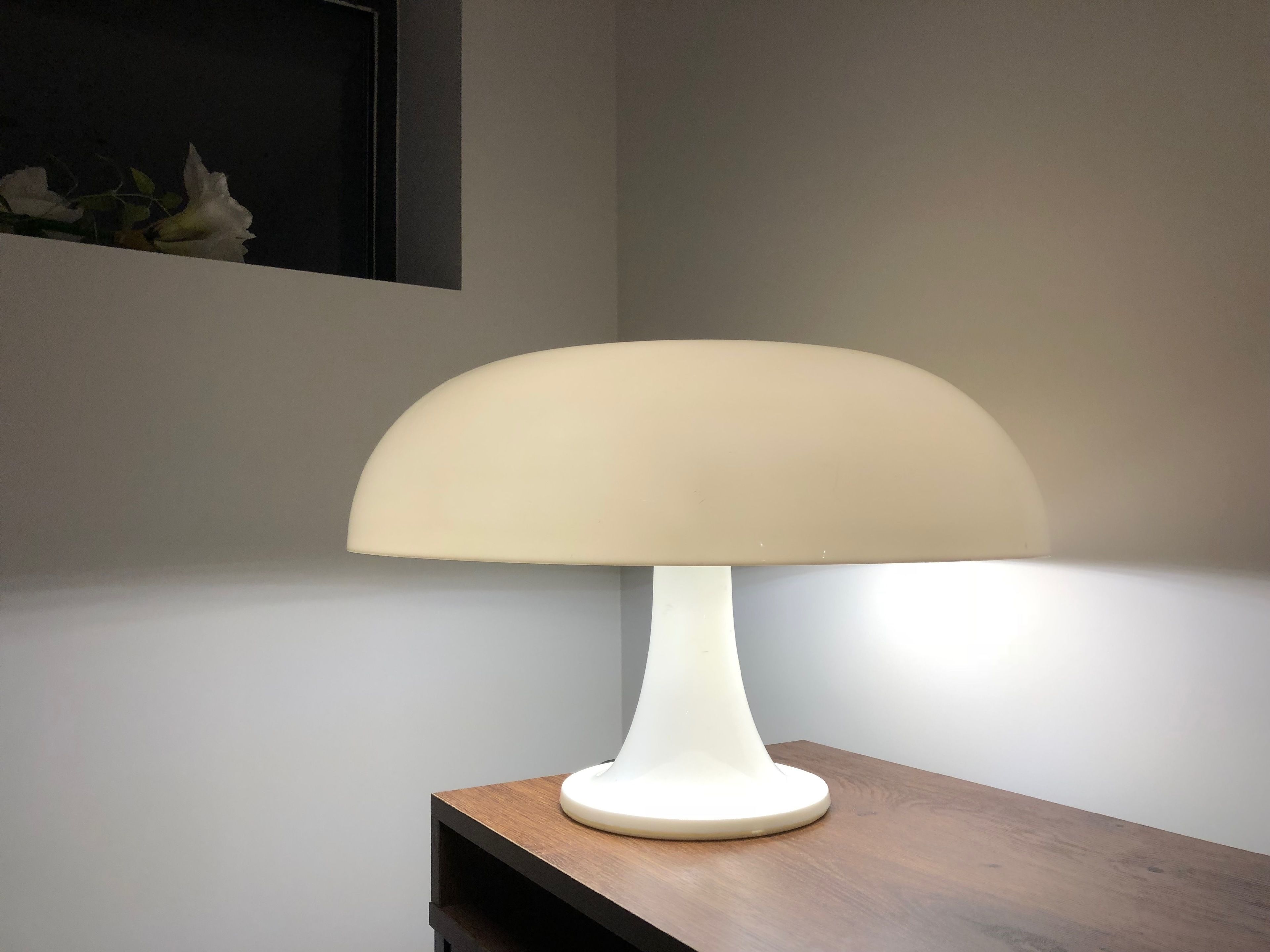 Een witte tafellamp met een koepelvormige lampenkap op een houten tafel, voor een effen muur.