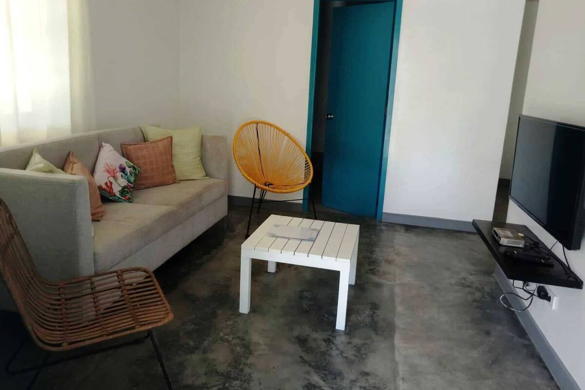 A living room with a sofa, a white coffee table, a wicker chair, a fan, a teal door, and a TV on a stand.