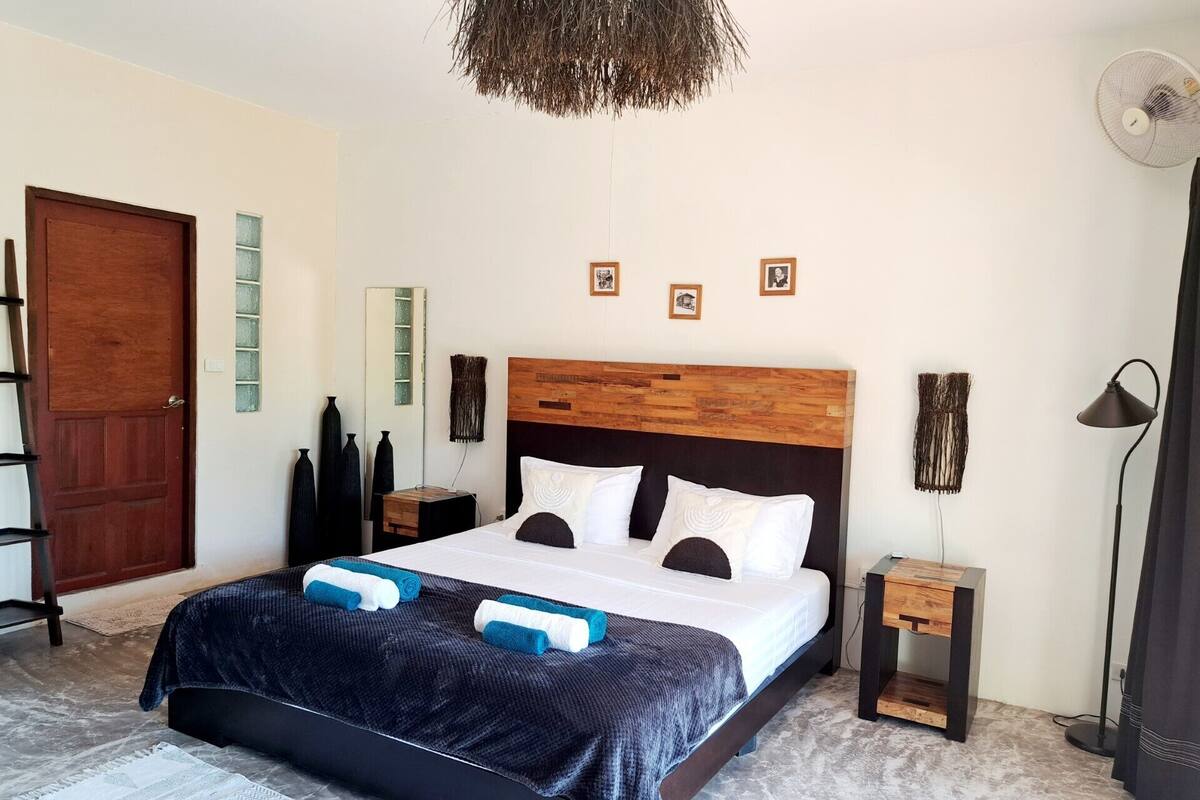 A modern bedroom with a wooden headboard, a large bed with white linens, two bedside tables, a standing fan, and a door with a glass panel.