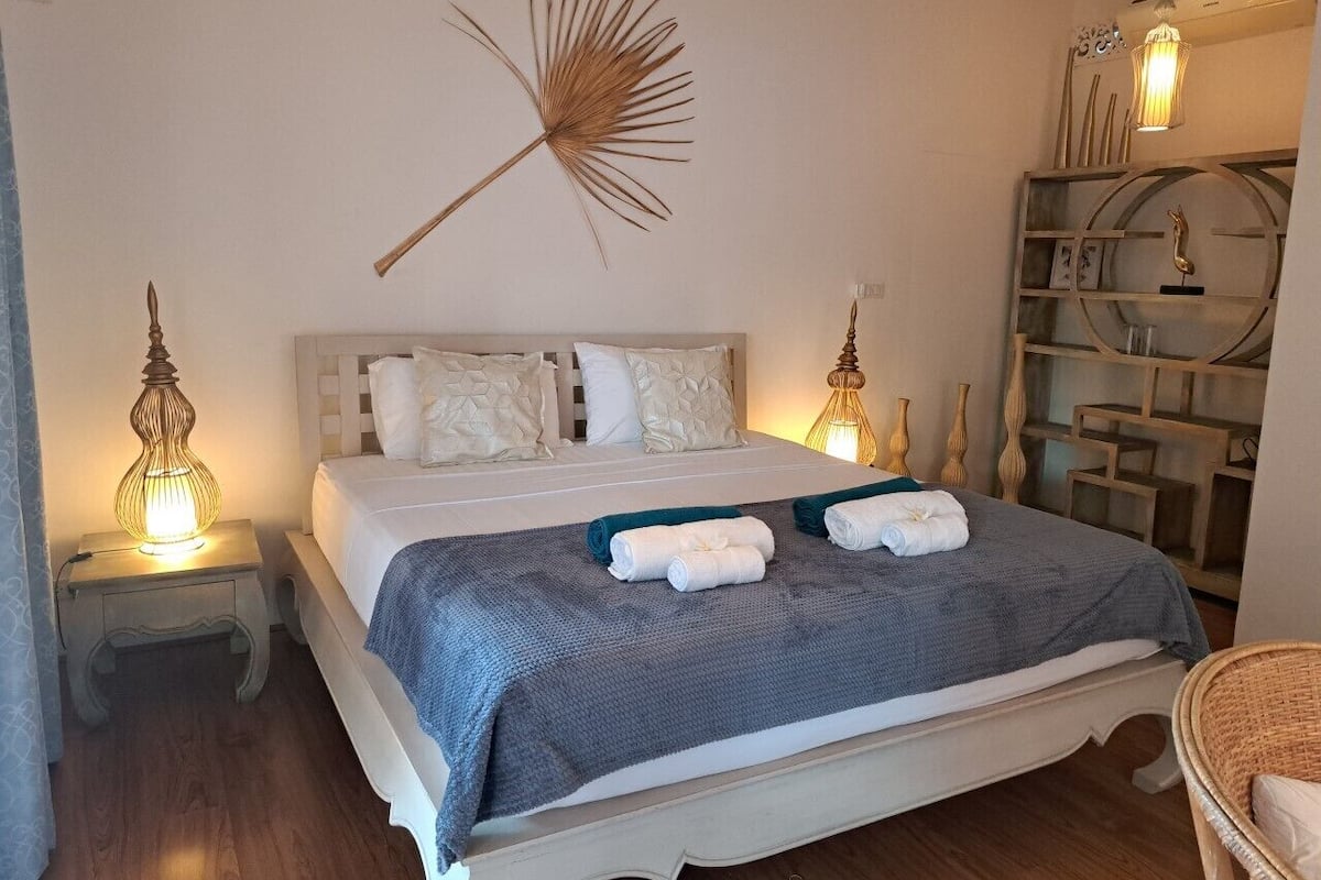 A neatly made bed with a blue blanket and two rolled towels, a wall-mounted fan, and a wooden side table with a lamp.