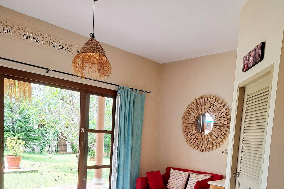 A bedroom with a bed, a red sofa, a small table, a hanging light fixture, and a view of the outdoors.