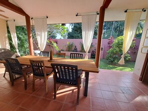 Un porche cubierto con una mesa y sillas de madera, rodeado de cortinas y vistas a un jardín con una estatua.