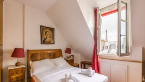 Une chambre à coucher avec un lit, deux tables de chevet, une commode, une fenêtre donnant sur des immeubles et un tableau accroché au mur.