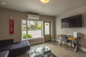 Una habitación moderna con sofá, una mesa de centro de vidrio, escritorio con silla, televisor de pantalla plana y un ventanal con vistas a la calle.