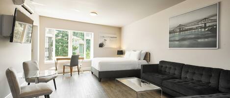 A modern hotel room with a bed, a dark sofa, a glass coffee table, a glass dining table with chairs, and a large window offering a view of greenery and a building.