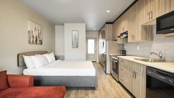 A modern hotel room with a bed, a sofa, a kitchen with wooden cabinets, a dishwasher, and a flat-screen TV.