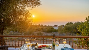 A table set with a bottle of wine, glasses, a book, and a plate of food on a balcony overlooking a sunset.