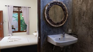 Ein Schlafzimmer mit Bett, einem Vorhangfenster und Blick auf Grünfläche. Das Badezimmer verfügt über ein Waschbecken, einen Spiegel und eine Steinwand.