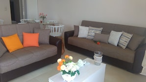 En stue med to sofaer, en med orange og en med hvide puder, et sofabord med blomsterarrangement og et lille bord med en vase blomster.