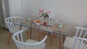 Et spiseområde med et glasbord omgivet af hvide stole. Der er en vase med blomster, en skål med snacks og to kopper på bordet.