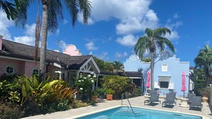Una piscina con sillas de descanso y sombrillas, rodeada de palmeras y un edificio con fachada rosa.