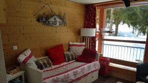 Ein gemütliches Blockhaus-Interieur mit einer Holzwand, einer Couch mit gemusterten Kissen, einem Tisch und einem Fenster mit Blick auf schneebedeckte Bäume.