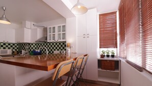 Cocina compacta con isla central, armarios blancos, salpicadero a cuadros y ventana con persianas.