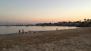 Una playa con arena, mar tranquilo y barcos al fondo.