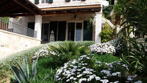 Un bâtiment en pierre doté d’un balcon, un jardin avec des fleurs blanches et un grand rocher au premier plan.