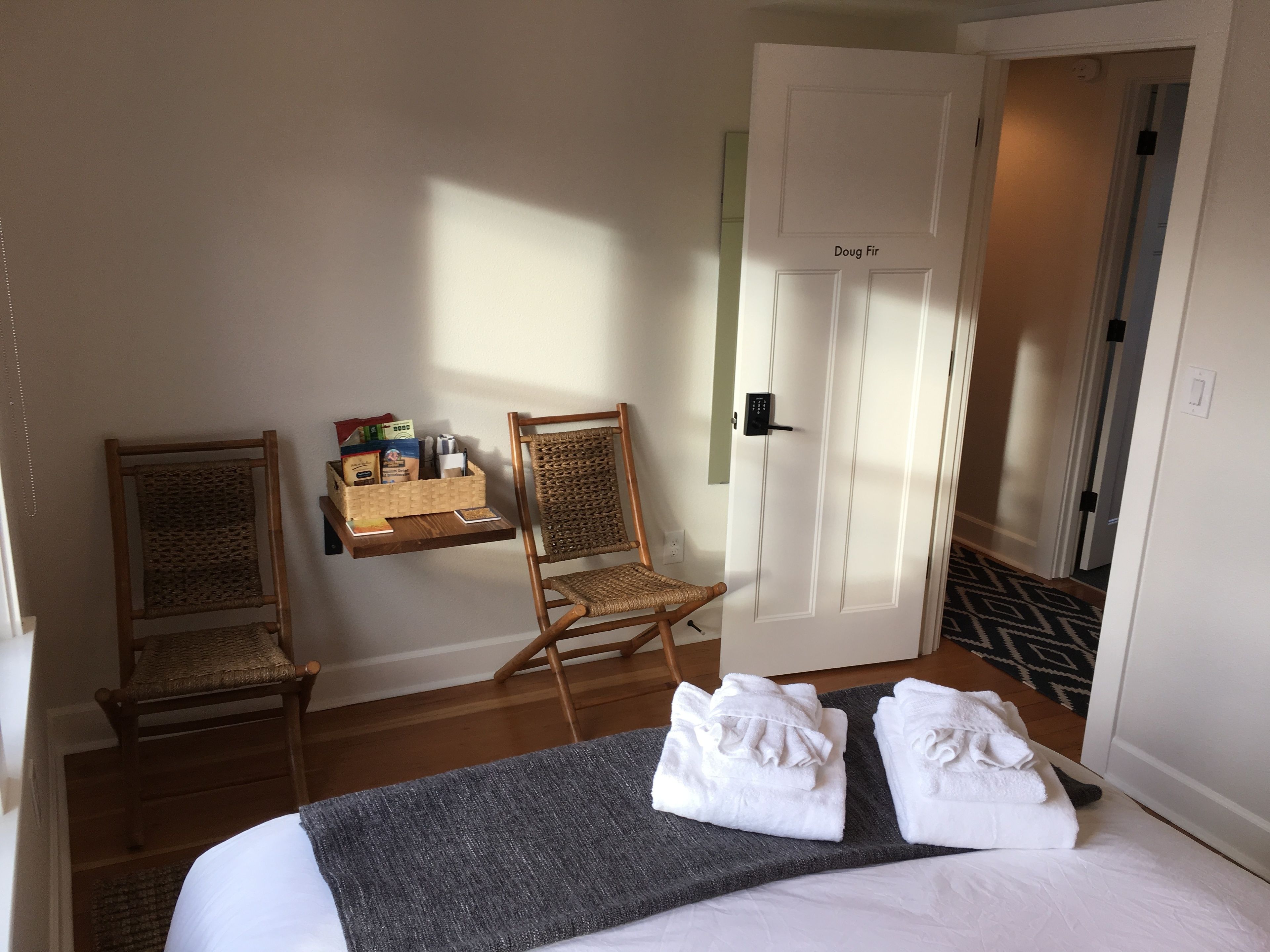Hotelkamer met een bed, twee rieten stoelen, een klein tafeltje met toiletartikelen en een deur met slot.