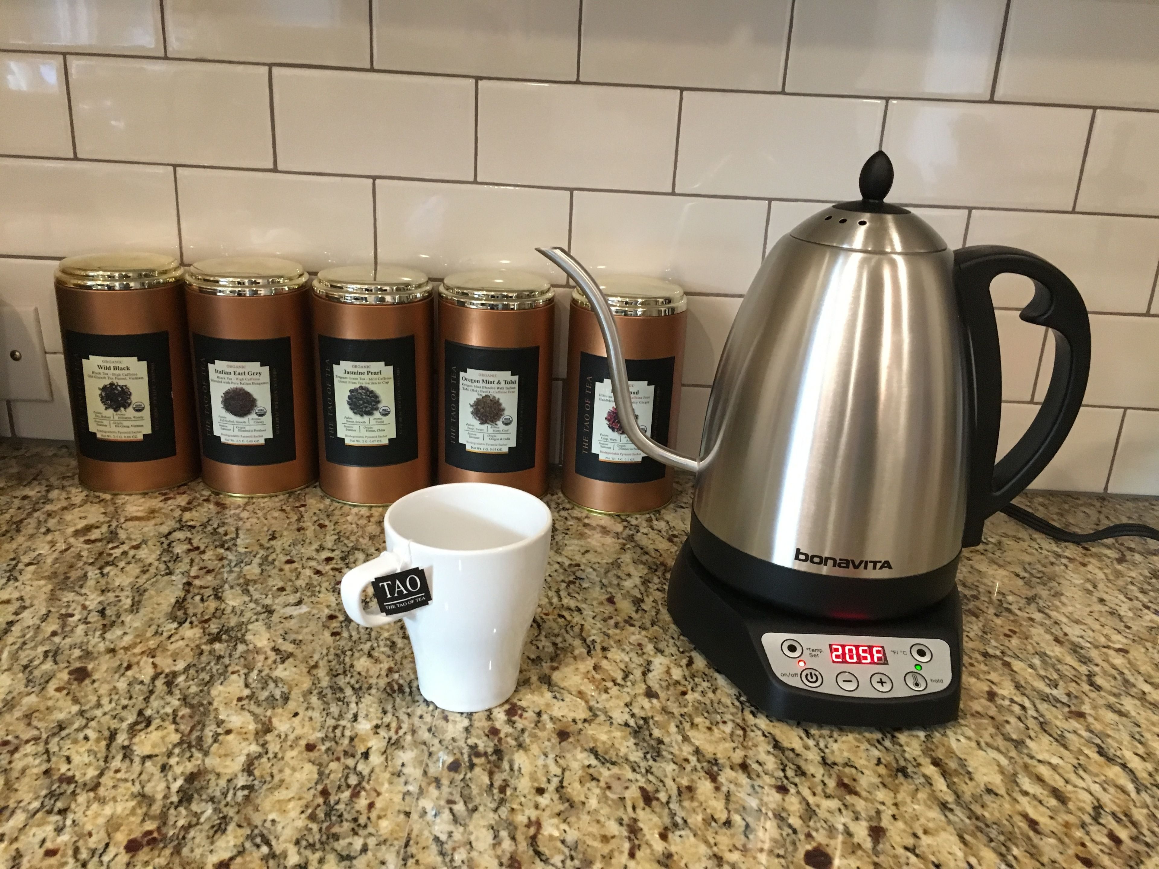 A kitchen with a stainless steel electric kettle and a white mug on a granite countertop. Behind the counter, there are four copper tea tins.
