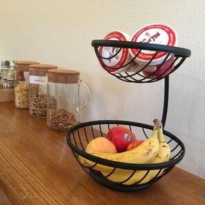 A kitchen counter with a fruit basket containing bananas, apples, and peaches. There are also glass jars with labels such as "Granola (GF)" and "Whole Grain." - The Alberta Petite Hotel (Portland)