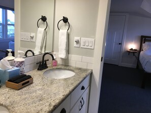 A hotel bathroom with a granite countertop, a white sink, a mirror, a towel rack, a nightstand with a lamp, and a bed with white bedding. - The Alberta Petite Hotel (Portland)