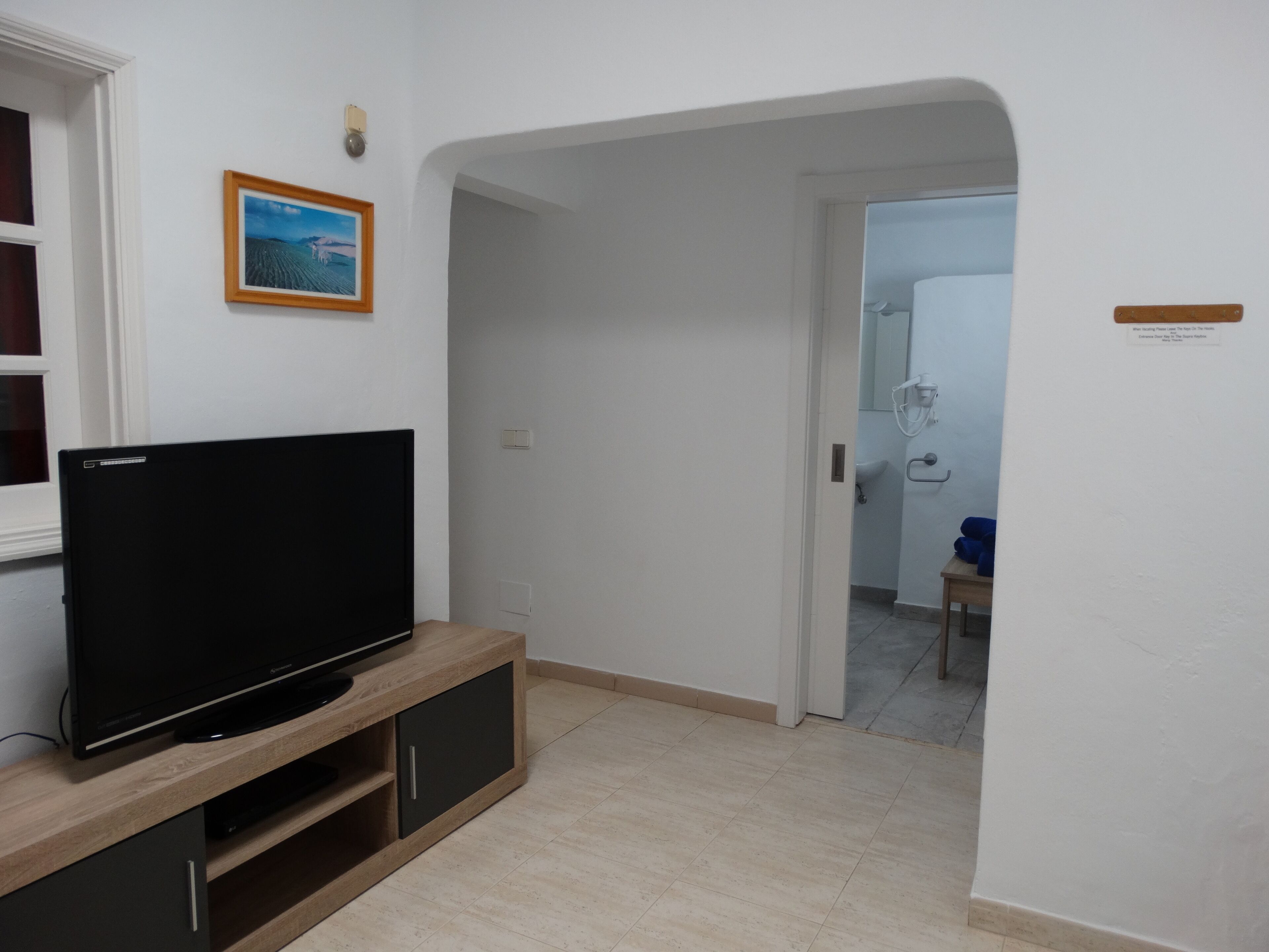 A modern living room with a flat-screen TV on a wooden stand, a framed picture on the wall, and a doorway leading to a bathroom.