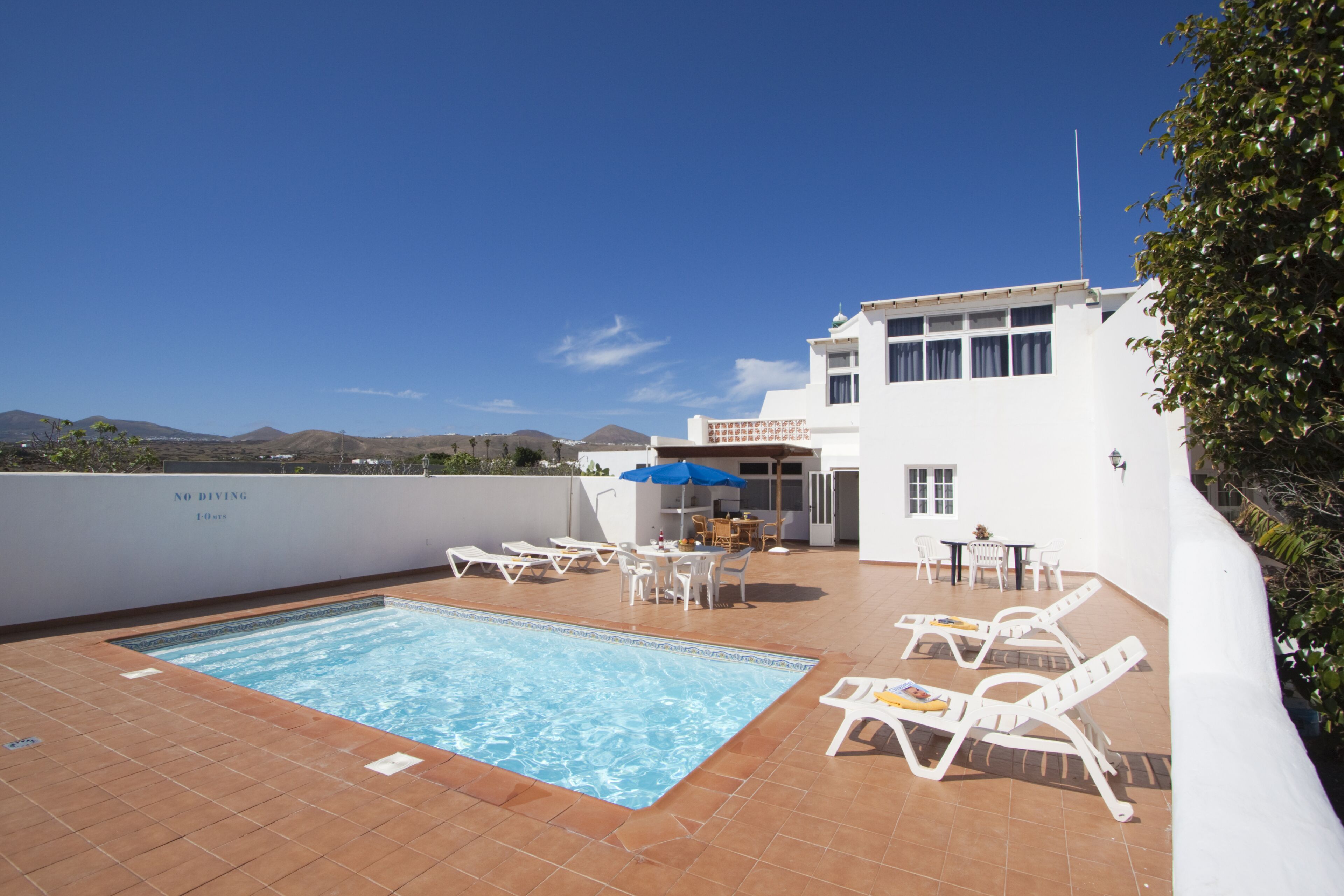 Zona de piscina en la azotea con sillones de descanso y una mesa de comedor, rodeada por un edificio blanco, un cielo azul despejado y colinas distantes al fondo.
