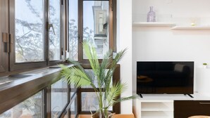 Een moderne woonkamer met een groot raam, een flatscreen televisie, een gevlochten plantenpot en een witte plank met decoratieve objecten.