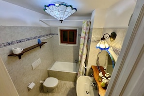Baño con inodoro, lavamanos y bañera. Hay un estante con artículos de aseo y una ventana pequeña.