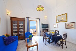 A living room with a blue sofa, a dining table with chairs, a television, and a view of the sea through a glass door.