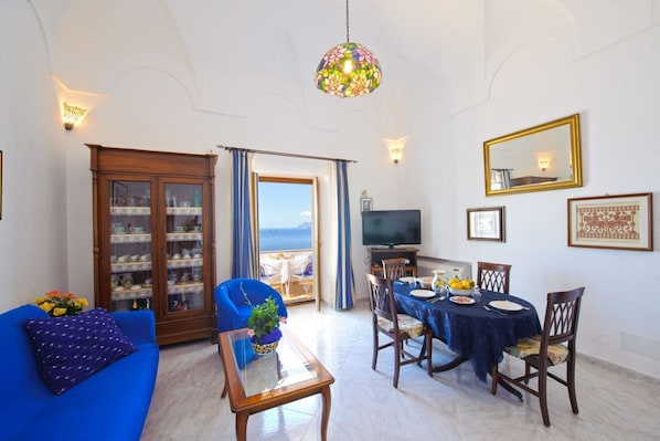 A living room with a blue sofa, a dining table with chairs, a television, and a view of the sea through a glass door.