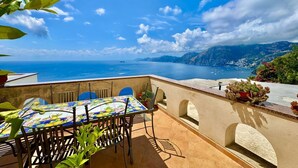 Eine Terrasse mit Tisch und Stühlen, mit Blick auf eine Küstenlandschaft mit Bergen und dem Meer.
