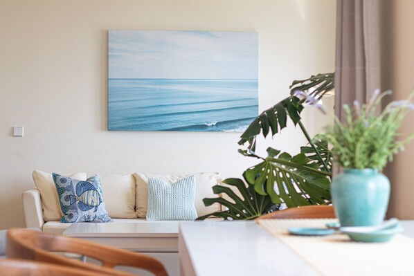 A living room with a sofa, a coffee table, and a painting of the ocean. - Gardenia Garden House with beautiful sea views, near the beach and the village. (Sternes)