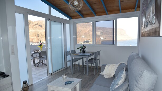 Ein modernes Wohnzimmer mit grauem Sofa, einem kleinen weißen Tisch und einem Essbereich mit Tisch und Stühlen. Große Fenster bieten Ausblick ins Freie.