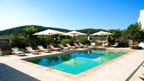 Piscina con sillas de descanso y sombrillas, rodeada de vegetación y colinas.