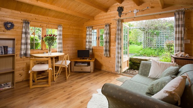 Interior de una cabaña de madera con mesa y sillas de madera, un sofá, un televisor y vistas al exterior.