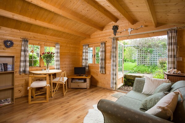 Interior de una cabaña de madera con mesa y sillas de madera, un sofá, un televisor y vistas al exterior.