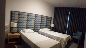 Habitación de hotel con dos camas individuales, una mesita de noche con lámpara, una silla y un amplio ventanal con cortinas.