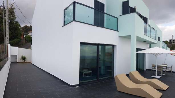 Un edificio moderno con techo plano, grandes ventanales y una terraza con dos sillones color beige.