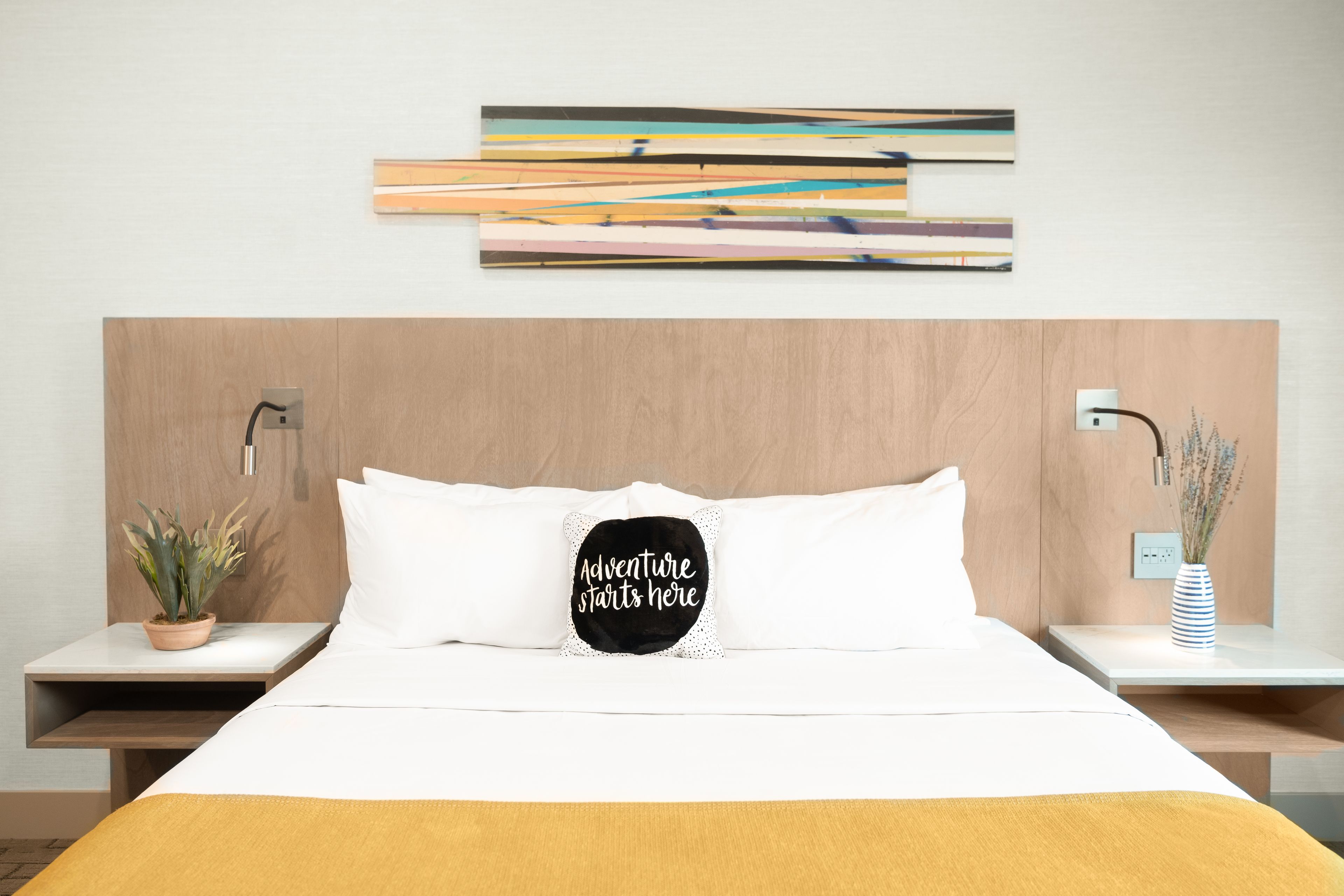 Een verzorgde hotelkamer met een bed, nachtkastje met een plant, een kunstwerk aan de muur en een decoratief kussen.