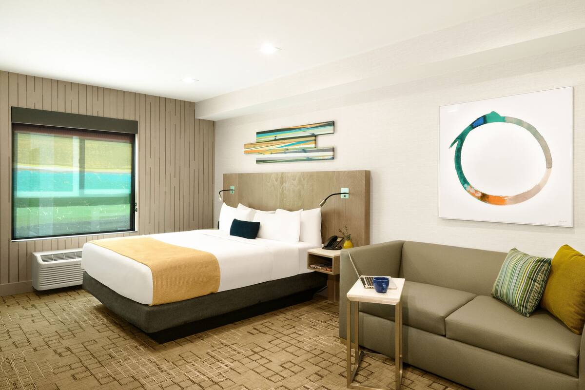 Une chambre d’hôtel avec un lit, un canapé, une fenêtre donnant sur un beau paysage, un tableau et une lampe.