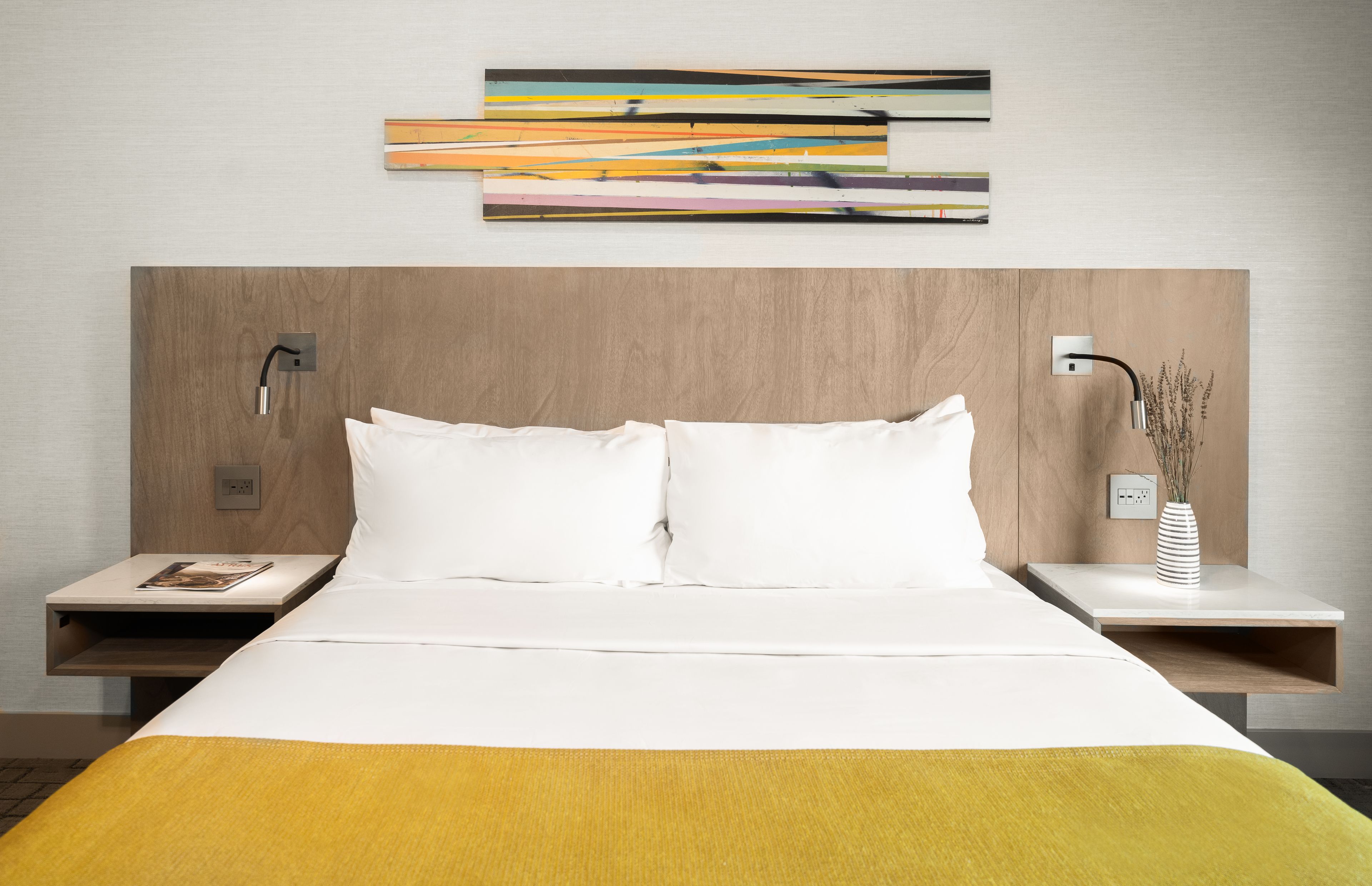 Habitación de hotel con una cama grande, dos mesitas de noche, una obra de arte abstracta moderna sobre la cama y un florero con plantas secas en la mesa de la derecha.