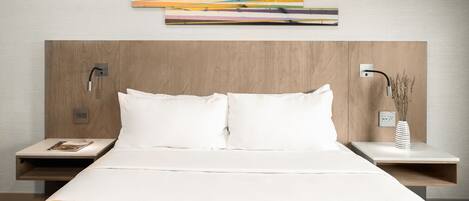 Habitación de hotel con una cama grande, dos mesitas de noche, una obra de arte abstracta moderna sobre la cama y un florero con plantas secas en la mesa de la derecha.