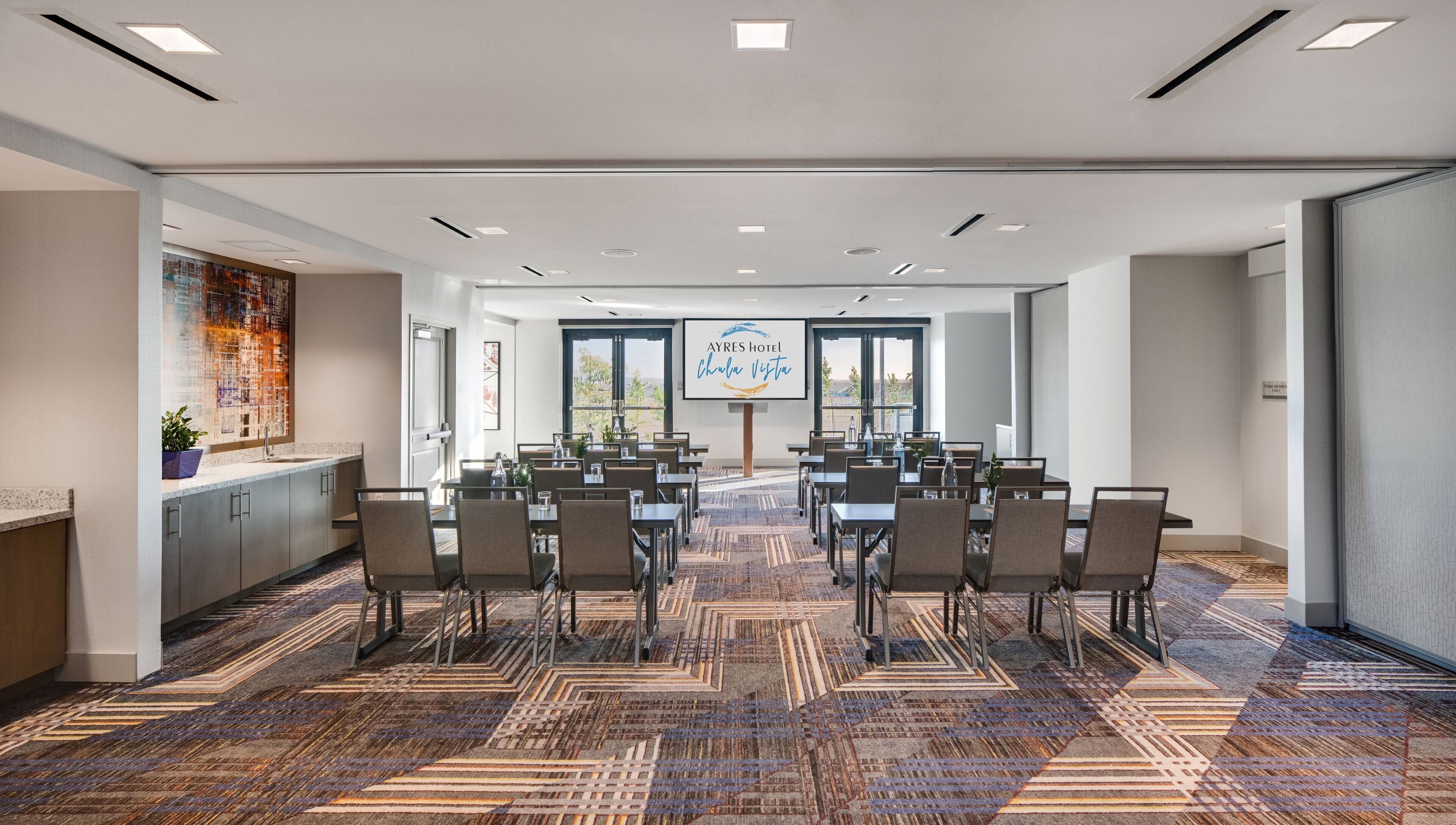 A conference room with a large screen, multiple tables, and chairs, carpeted floor, and a view of the outdoors.