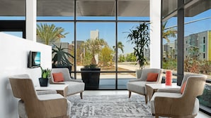 A modern lounge area with two grey upholstered chairs, a small table, and a large window offering a view of palm trees and buildings.