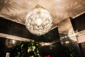A modern chandelier with a geometric design, a dark wall with a reflective surface, and a potted plant in the foreground. - Awesome Hotel (Jeonju)
