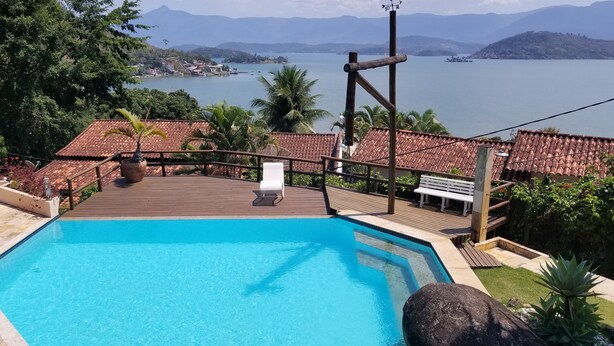A pool with a wooden deck, a white lounge chair, and a view of a lake and mountains. - Vista paradisiaca (Angra dos Reis)