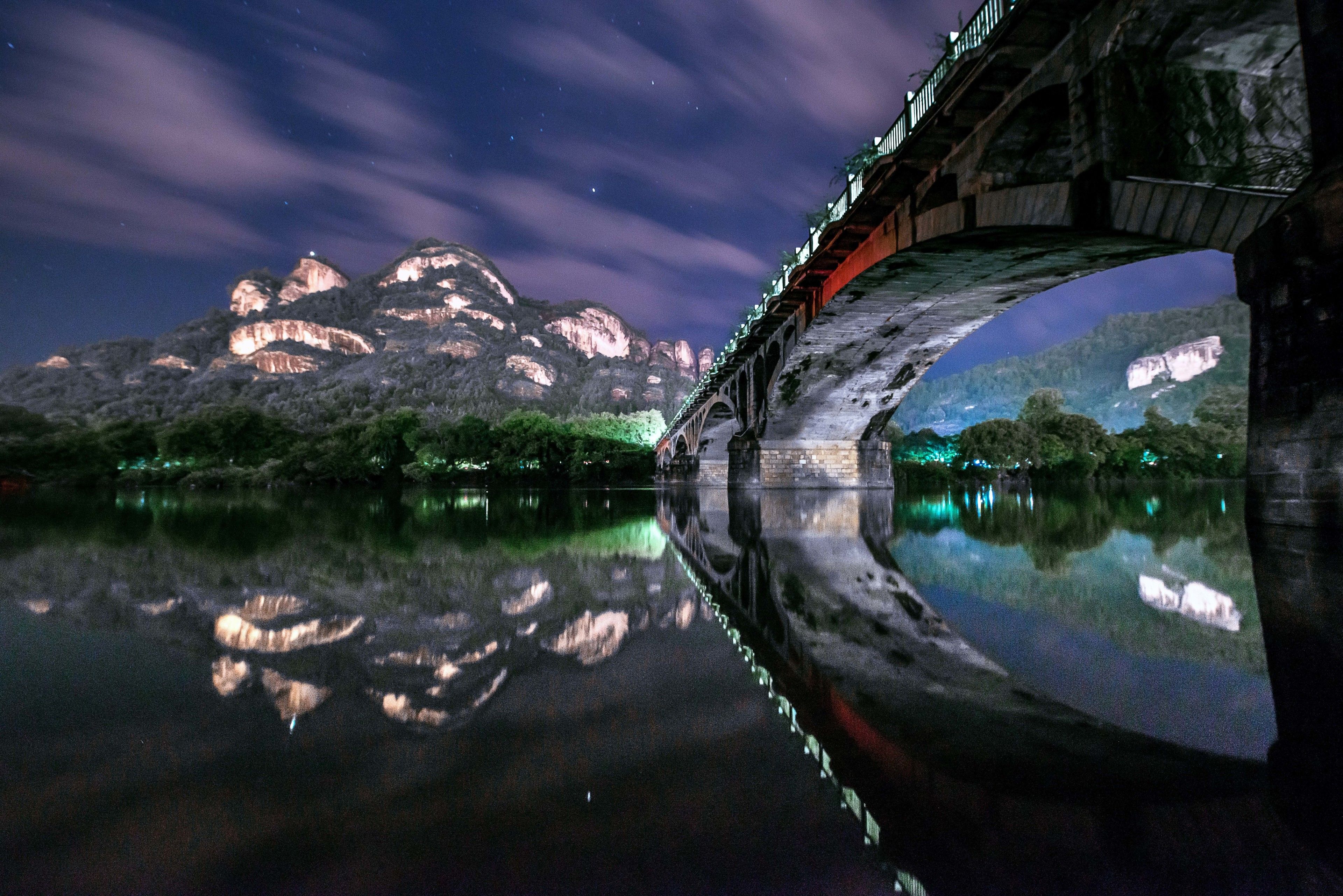 Un pont se reflétant dans l’eau calme, entouré de montagnes et surplombant un ciel étoilé.