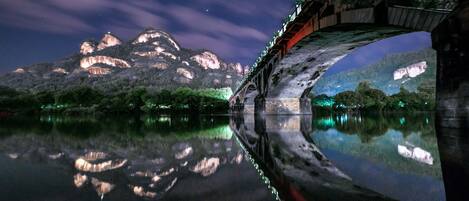 Un pont se reflétant dans l’eau calme, entouré de montagnes et surplombant un ciel étoilé.